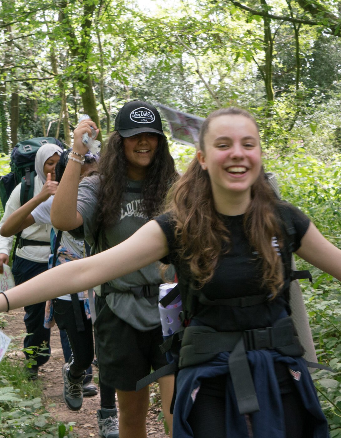 A group of young people hiking through a lush, green forest, all wearing backpacks. The person at the front, a smiling girl with long brown hair, has her arms spread wide in excitement, holding a red item in one hand and a map in the other. Behind her, two other participants follow, also smiling, one wearing a cap and the other a hoodie. Sunlight filters through the trees above.