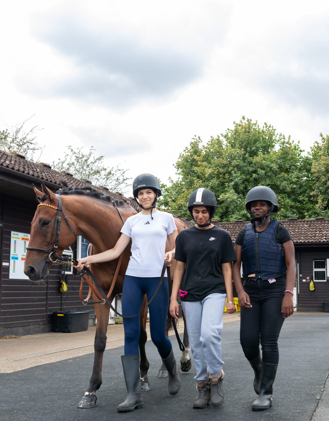 Three individuals are walking together with a horse in an outdoor stable area. The first person, wearing a white shirt and riding pants, is leading the horse. The second person, dressed in a black shirt and light pants, is walking alongside, while the third person, wearing a black vest and helmet, is also accompanying them. The background features stable buildings and greenery under a cloudy sky.