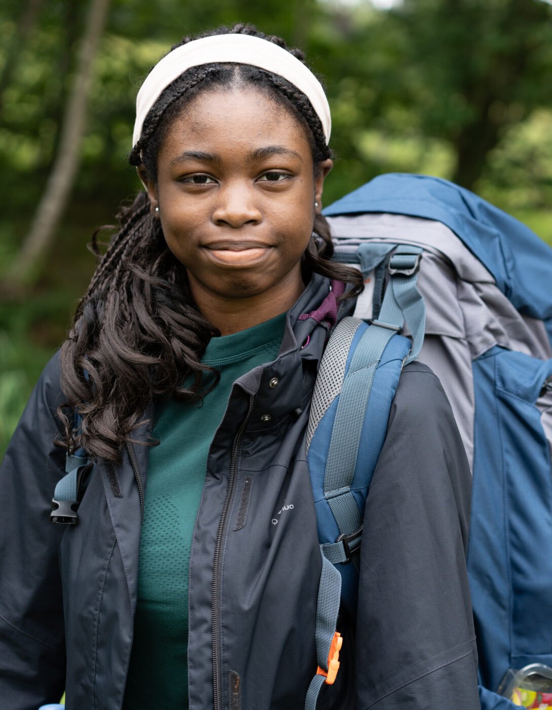 A young woman with long, dark hair wearing a black jacket and a white headband stands outdoors, smiling slightly. She has a large backpack secured to her back, which includes an orange sleeping bag. The background features greenery and trees, suggesting a natural setting.