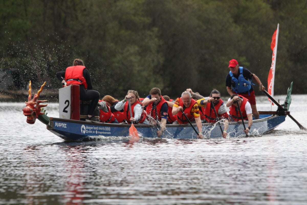 A team of people in a dragon boat paddling vigorously on a calm body of water. All team members are wearing red life jackets, and the boat features a dragon-shaped design on the front. Splashes of water surround the paddlers as they work in unison, with a helmsman at the back steering and shouting commands. Trees and greenery line the background, creating a natural setting.