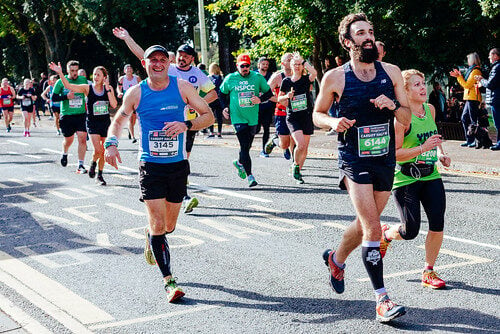 A group of runners participating in a race on a sunny day. The runners wear numbered race bibs, with some displaying logos for charities like NSPCC. One runner in a blue tank top smiles and waves at the camera, while others focus on running. The background shows cheering spectators and trees, creating a lively atmosphere.