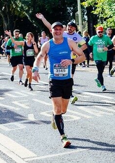 A group of runners participating in a race on a sunny day. The runners wear numbered race bibs, with some displaying logos for charities like NSPCC. One runner in a blue tank top smiles and waves at the camera, while others focus on running. The background shows cheering spectators and trees, creating a lively atmosphere.