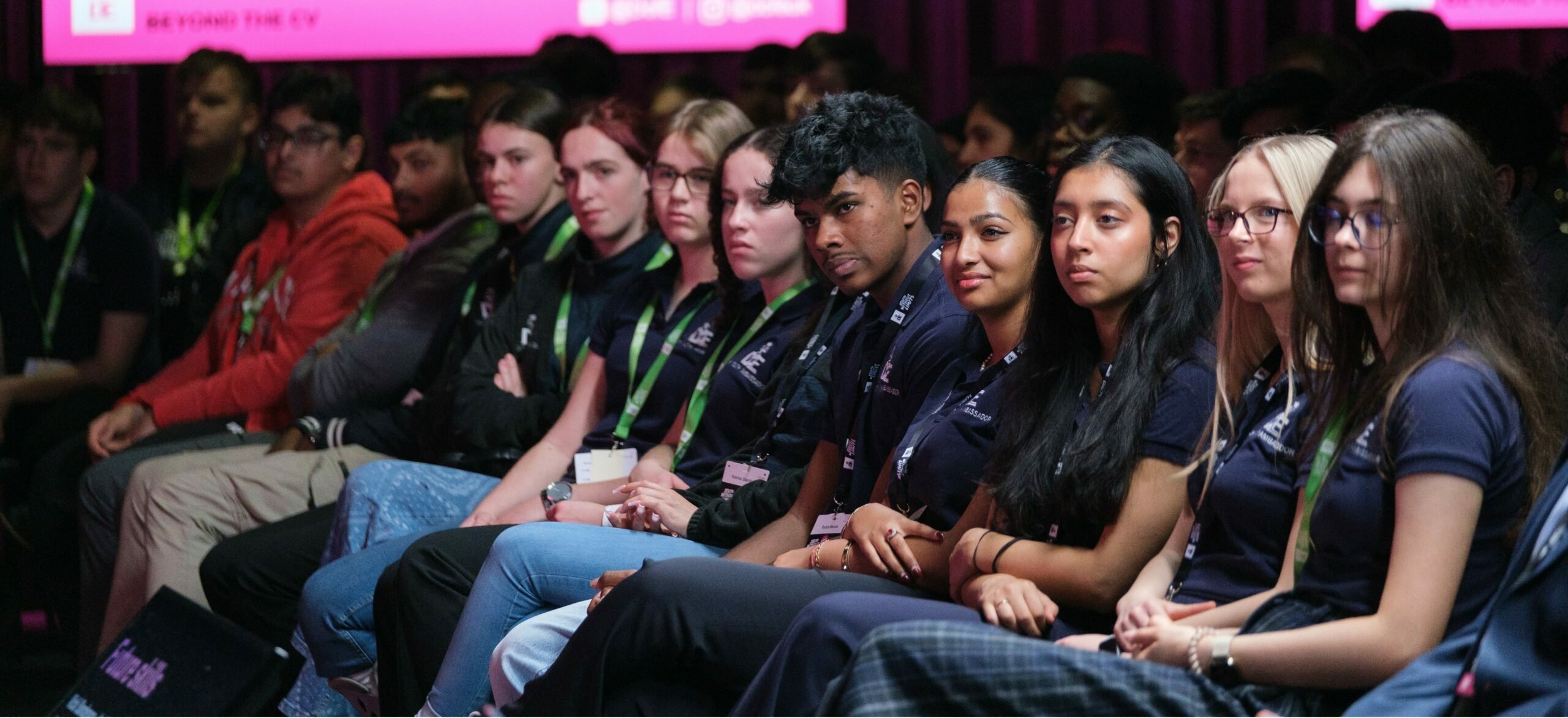 A diverse group of young people sitting in an audience, attentively listening to a presentation. They are wearing matching dark shirts and have green lanyards around their necks. The background features a stage with pink and purple lighting, displaying the text 