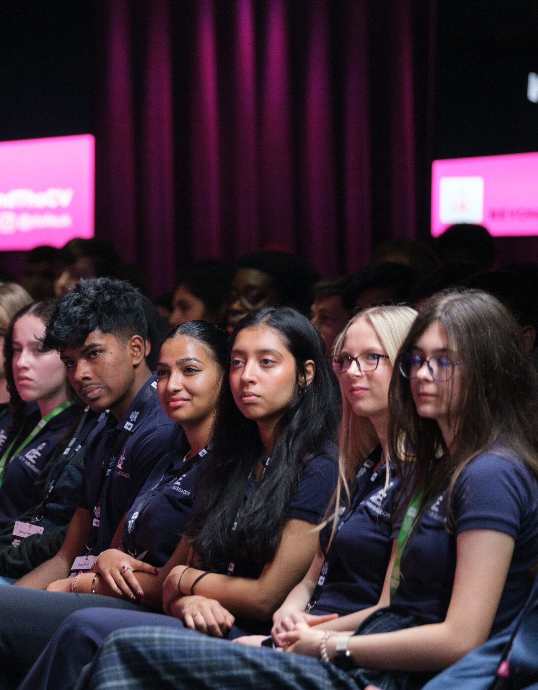 A diverse group of young people sitting in an audience, attentively listening to a presentation. They are wearing matching dark shirts and have green lanyards around their necks. The background features a stage with pink and purple lighting, displaying the text 