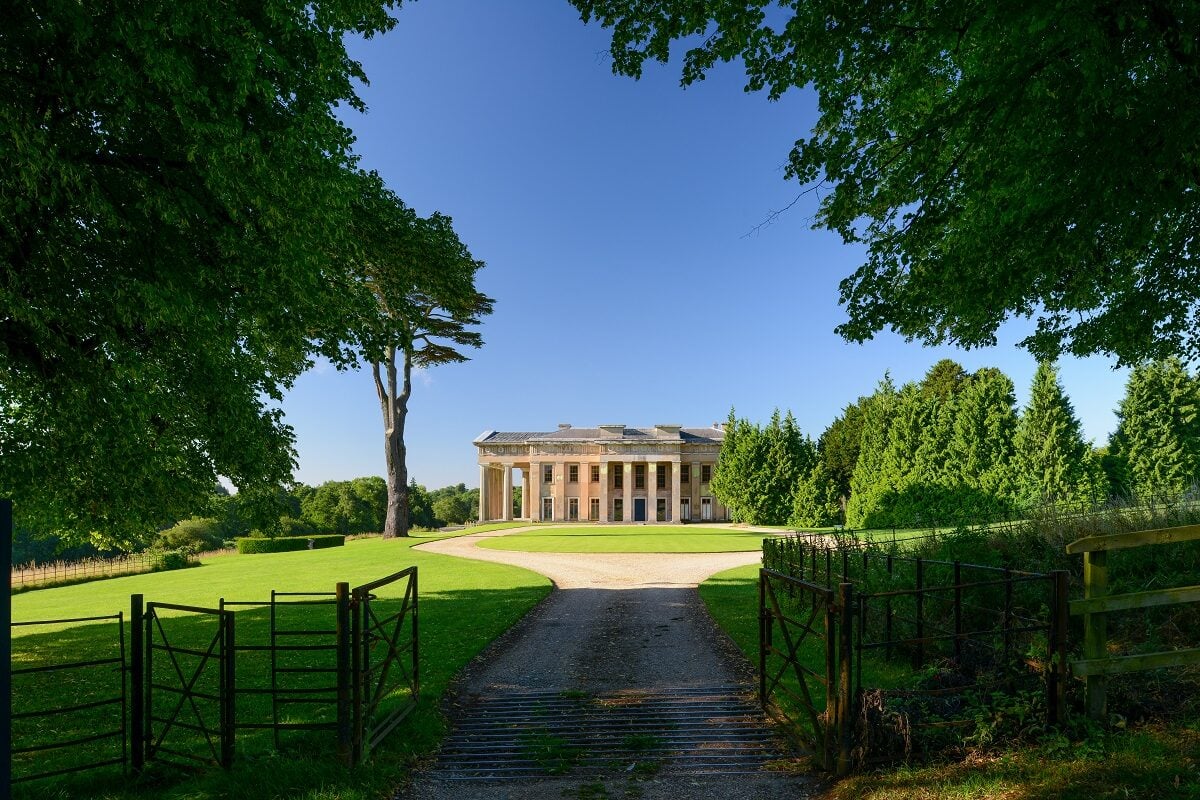 A grand neoclassical mansion with tall columns and a symmetrical facade, surrounded by a well-maintained lawn and lush greenery. The building is viewed from a gated entrance on a sunny day, with a bright blue sky and trees framing the scene.