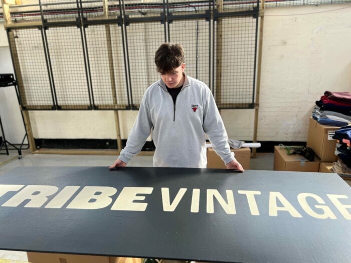 Freddie wearing a light grey sweatshirt with a small logo on the chest is standing indoors, looking down at a large black sign that reads 'TRIBE VINTAGE' in bold white letters. The setting appears to be a workspace, with cardboard boxes and folded clothing visible in the background.