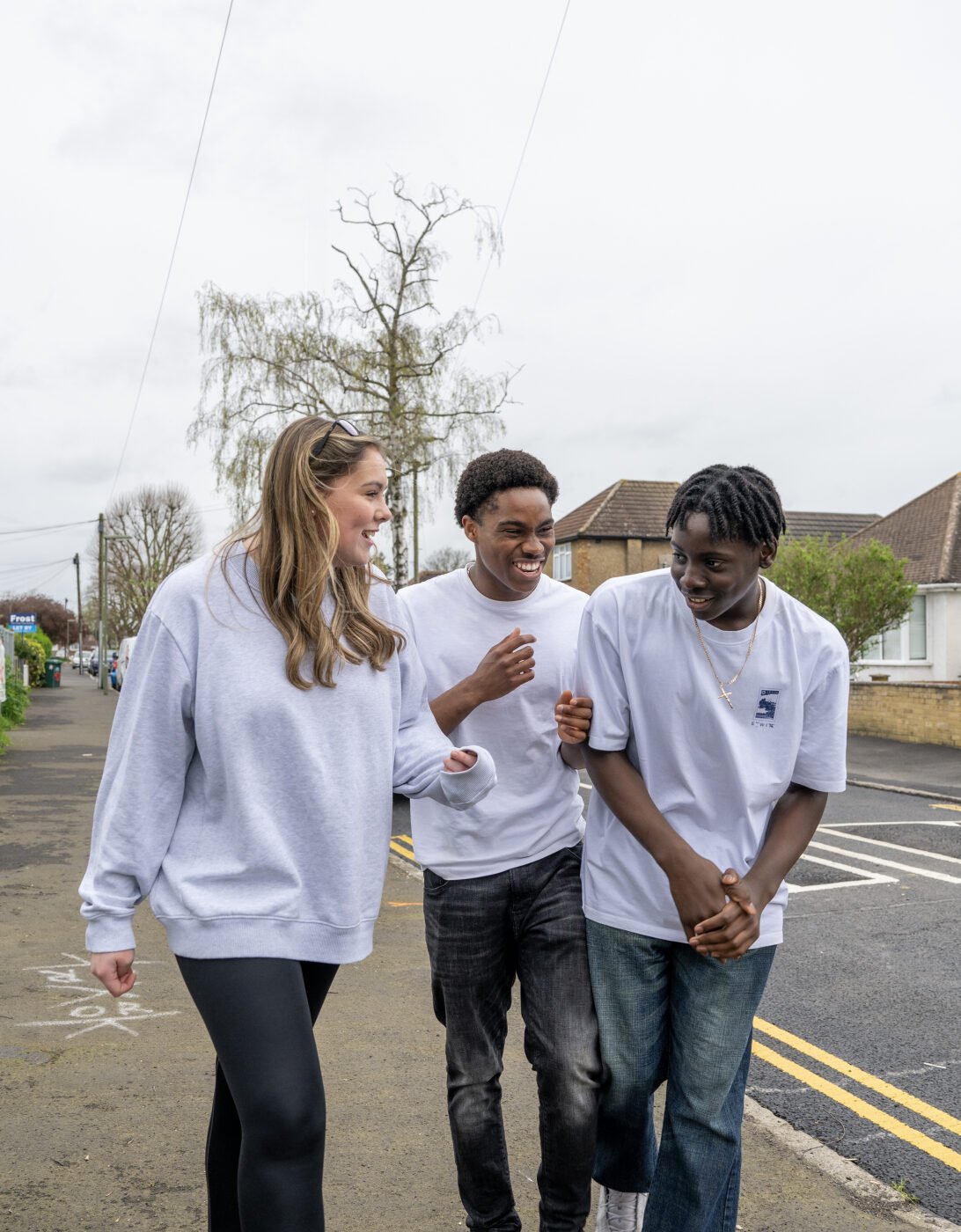 Three teenagers walking together and laughing on a suburban street. A girl in a grey sweatshirt is chatting with two boys, one wearing a white t-shirt and jeans, and the other in a white t-shirt and dark trousers. They appear to be enjoying each other's company on a cloudy day, with houses and a metal fence in the background.