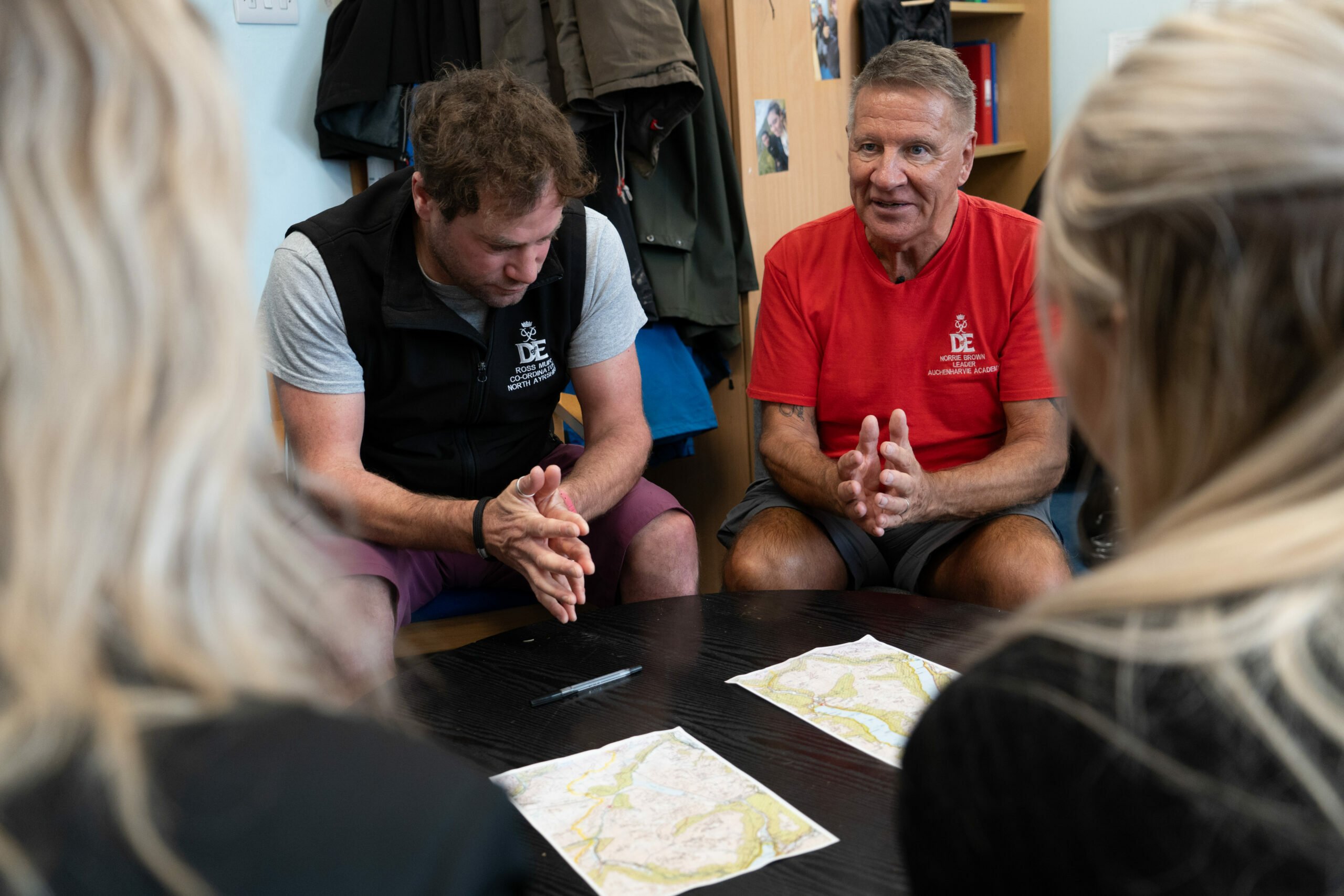 Two DofE leaders sit at a table, engaged in discussion with a group of young participants, as they review maps spread on the table. One leader wears a black DofE-branded vest, and the other wears a red DofE-branded t-shirt. The participants are partially visible, seen from behind, with their focus on the leaders. The setting is indoors, with coats and shelves visible in the background.