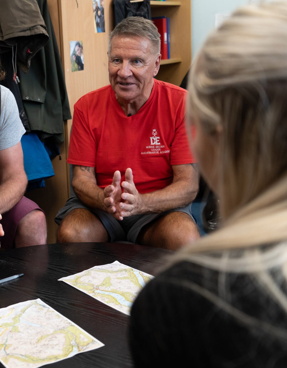 Two DofE leaders sit at a table, engaged in discussion with a group of young participants, as they review maps spread on the table. One leader wears a black DofE-branded vest, and the other wears a red DofE-branded t-shirt. The participants are partially visible, seen from behind, with their focus on the leaders. The setting is indoors, with coats and shelves visible in the background.