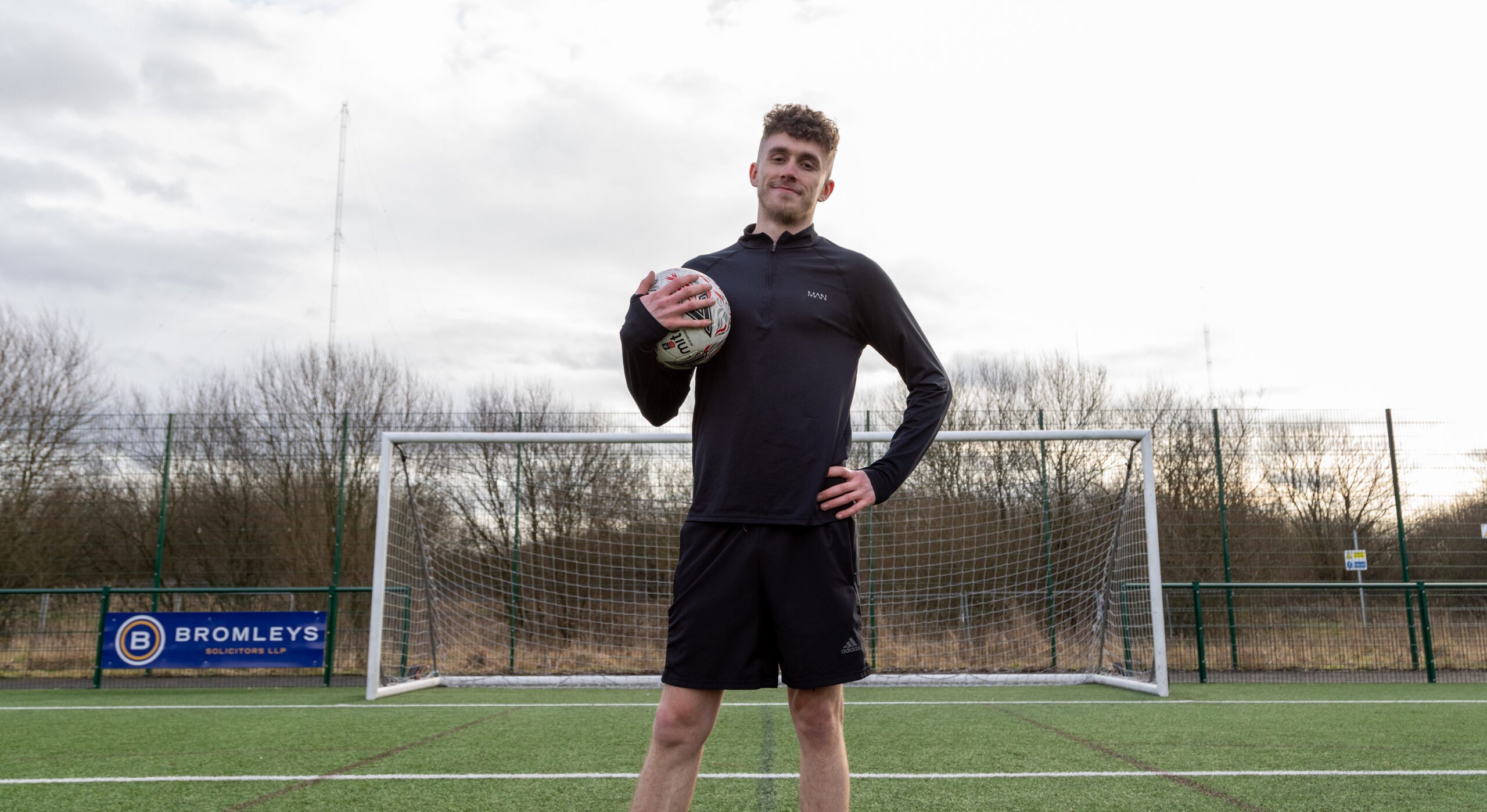 Jack is standing confidently on a football field in front of a goalpost, holding a football under one arm. He is wearing a black long-sleeve athletic top and black shorts, with a faint smile on his face. The background shows a fenced area with leafless trees and a cloudy sky.