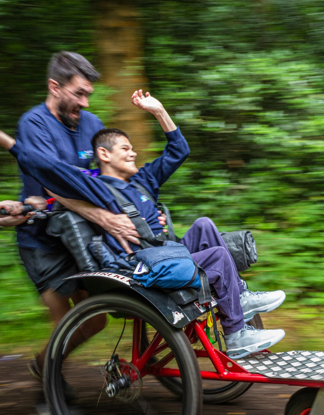 A boy with a joyful expression is being pushed in a specialized wheelchair along a wooded path. He is wearing a blue shirt and has his arms raised in excitement. Two adults, one in the foreground and one behind, are helping to push the wheelchair, both smiling and enjoying the moment. The background is a blur of green foliage, suggesting movement and speed.