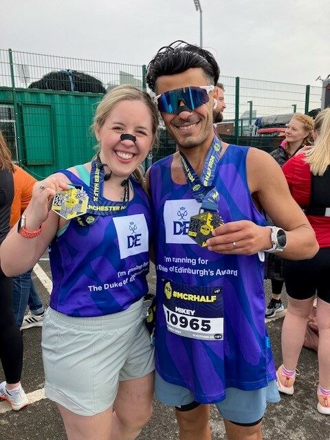 wo runners smiling and holding their medals after completing a race. Both are wearing purple Duke of Edinburgh's Award running tops. The woman on the left has light hair tied back, is wearing light-colored shorts, and has face paint resembling a dog's nose and whiskers. The man on the right is wearing reflective sunglasses and blue shorts, with 'Mikey' and '10965' visible on his race bib. A crowd and race fencing are visible in the background.