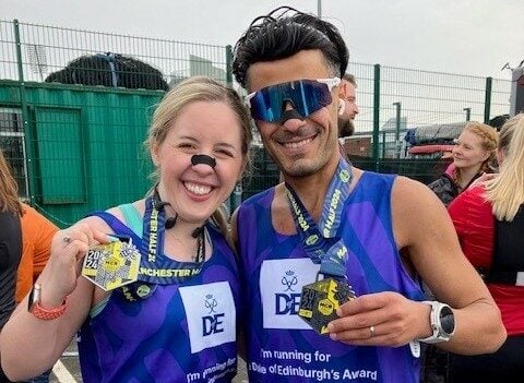 wo runners smiling and holding their medals after completing a race. Both are wearing purple Duke of Edinburgh's Award running tops. The woman on the left has light hair tied back, is wearing light-colored shorts, and has face paint resembling a dog's nose and whiskers. The man on the right is wearing reflective sunglasses and blue shorts, with 'Mikey' and '10965' visible on his race bib. A crowd and race fencing are visible in the background.