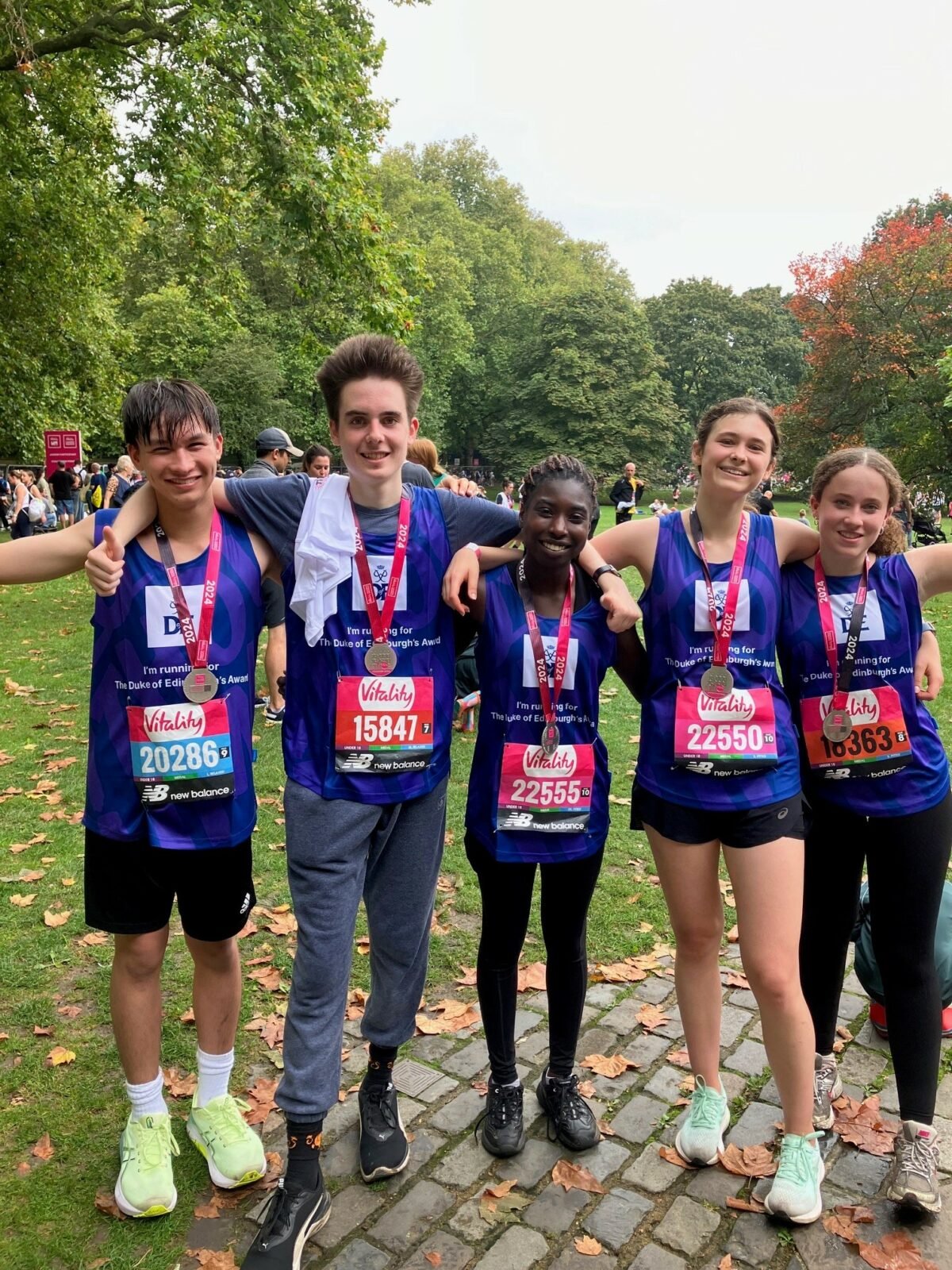 A group of five young runners stands together in a park, smiling and wearing medals around their necks after completing a race. They are dressed in purple running vests with “The Duke of Edinburgh’s Award” branding and have their race numbers visible on their chests. The background shows trees, grass, and other participants in the event, suggesting a lively and celebratory atmosphere.