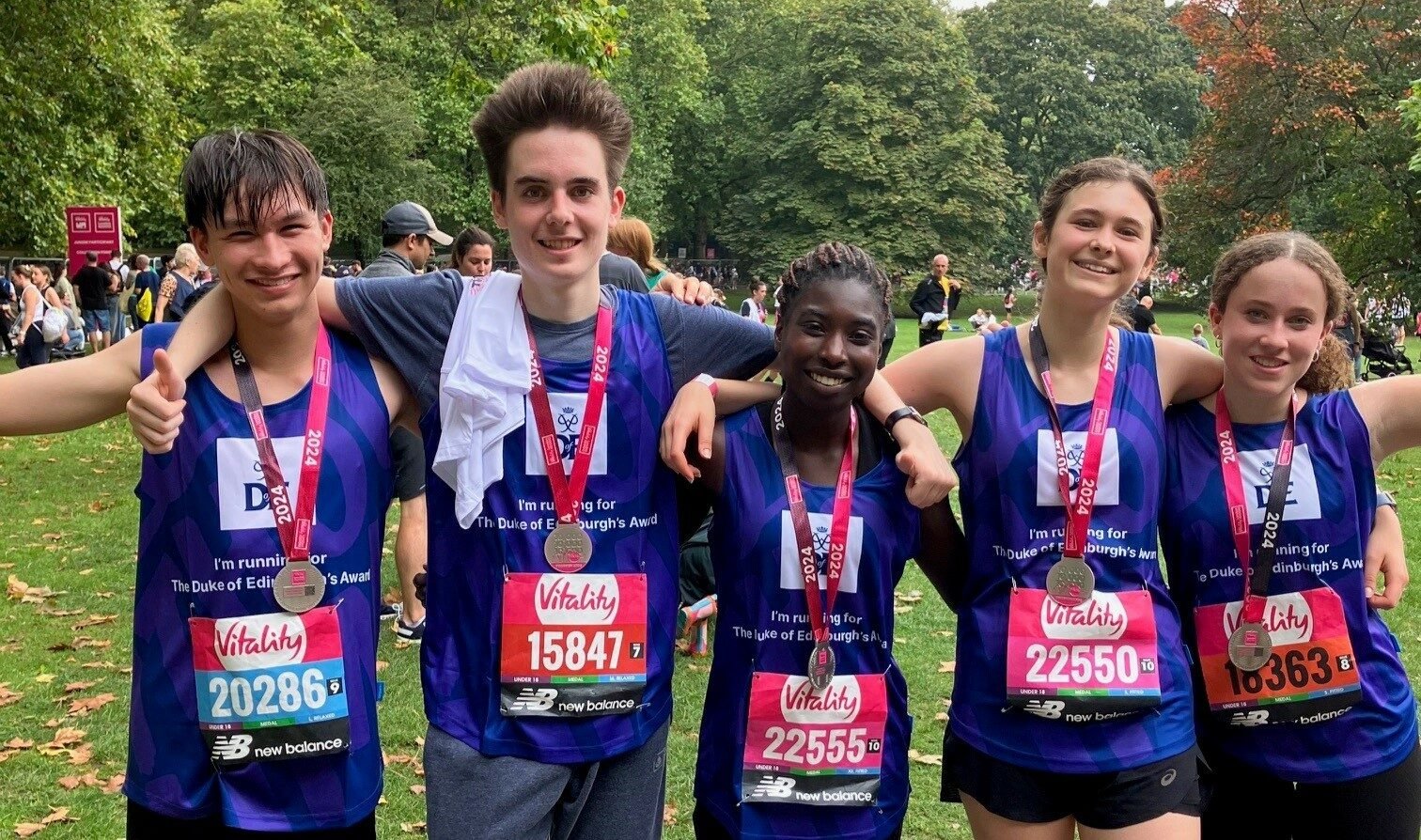 A group of five young runners stands together in a park, smiling and wearing medals around their necks after completing a race. They are dressed in purple running vests with “The Duke of Edinburgh’s Award” branding and have their race numbers visible on their chests. The background shows trees, grass, and other participants in the event, suggesting a lively and celebratory atmosphere.