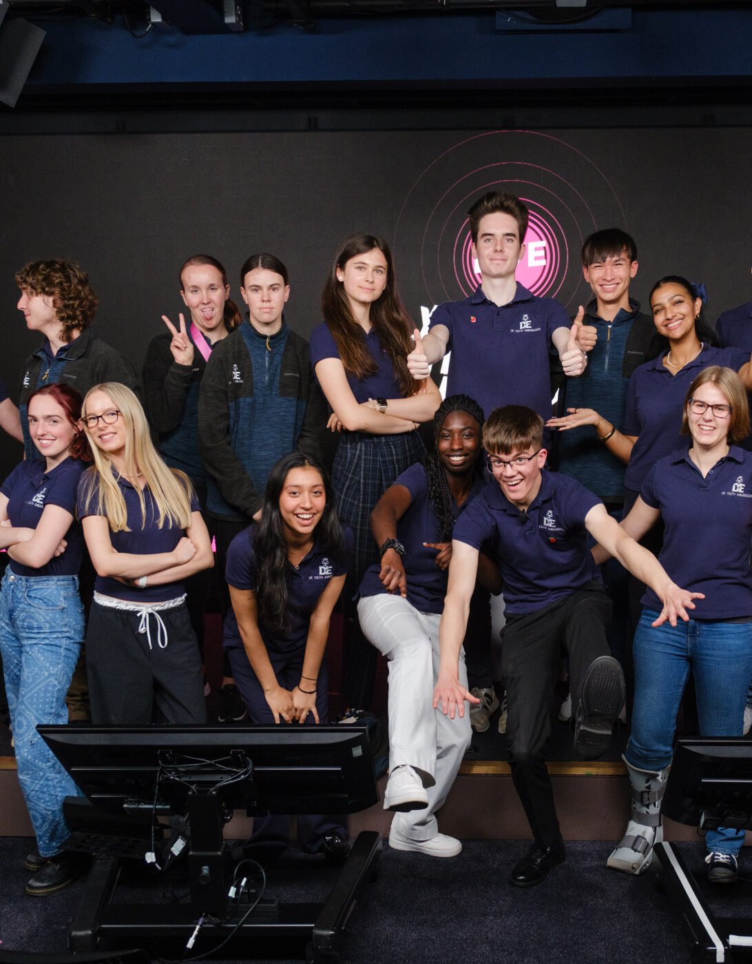 A diverse group of young people poses together in a modern event space, smiling and making playful gestures. They are wearing matching navy blue shirts and are arranged in a dynamic formation, with some individuals standing and others kneeling or sitting. The background features a large screen with the text 