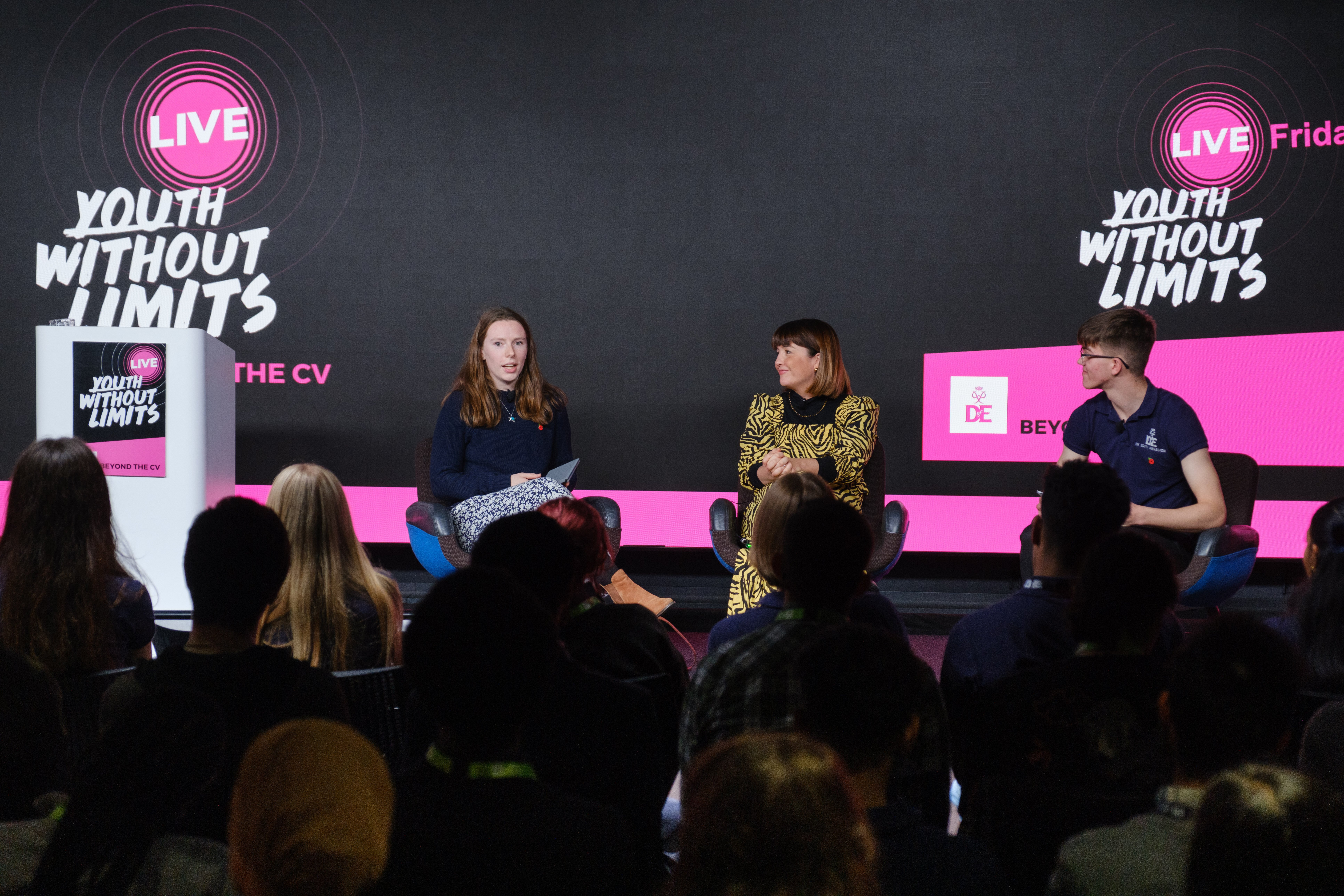 A panel discussion taking place on stage, featuring three speakers. One woman is seated on the left, holding notes and speaking. The second woman, dressed in a patterned dress, is in the center, listening attentively. A man in a blue shirt is seated on the right. The audience is visible in the foreground, facing the speakers. The backdrop displays the text 