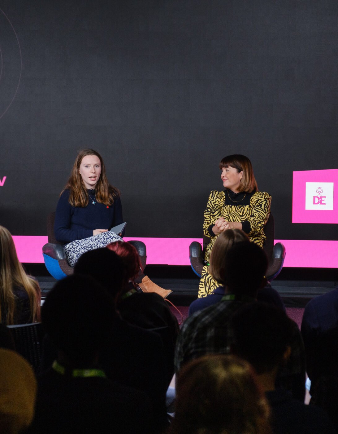 A panel discussion taking place on stage, featuring three speakers. One woman is seated on the left, holding notes and speaking. The second woman, dressed in a patterned dress, is in the center, listening attentively. A man in a blue shirt is seated on the right. The audience is visible in the foreground, facing the speakers. The backdrop displays the text 