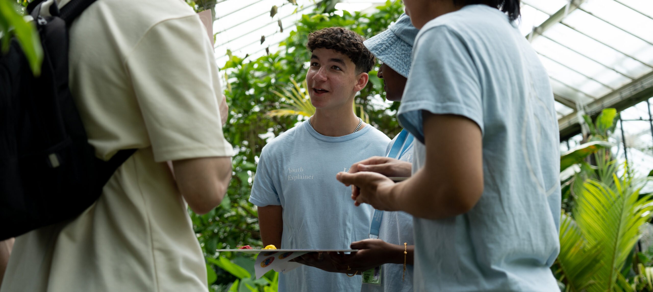 A young person wearing a light blue shirt labeled 'Youth Explainer' speaks enthusiastically to a small group of people in a greenhouse, surrounded by lush green plants. The group appears to be engaged in conversation, with one holding a clipboard or folder.