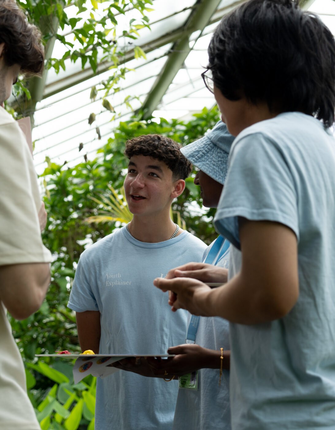 A young person wearing a light blue shirt labeled 'Youth Explainer' speaks enthusiastically to a small group of people in a greenhouse, surrounded by lush green plants. The group appears to be engaged in conversation, with one holding a clipboard or folder.