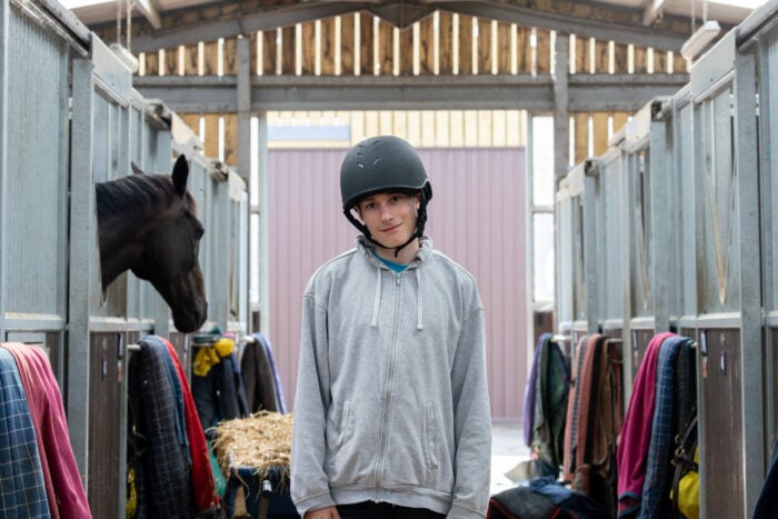 Ethan-Thomas wearing a helmet stands in a stable, surrounded by horse stalls. One horse's head is visible in the stall to the left. The stalls are lined with colorful blankets and hay, and the stable has wooden beams and a bright background. The person has a relaxed expression and is dressed in casual clothing.