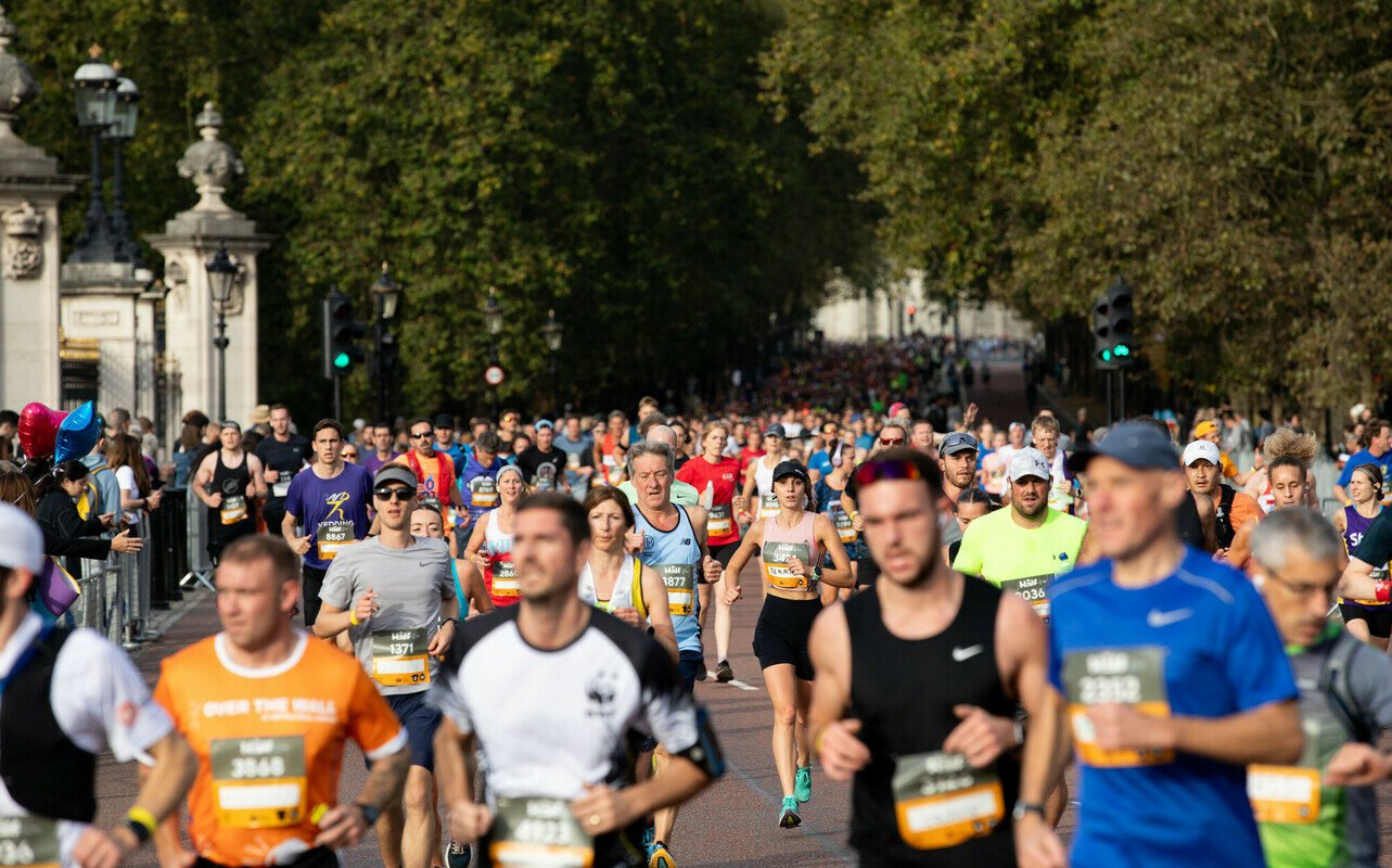 Marathon runners in athletic attire participate in a race on a wide, tree-lined road. The image captures a large crowd of participants, with some in the foreground wearing numbered bibs and determined expressions. A mix of men and women are visible, running at different paces, while spectators with balloons line the edge of the racecourse. The background showcases lush green trees and historic stone architecture under a clear sky, suggesting a scenic and energetic outdoor event.