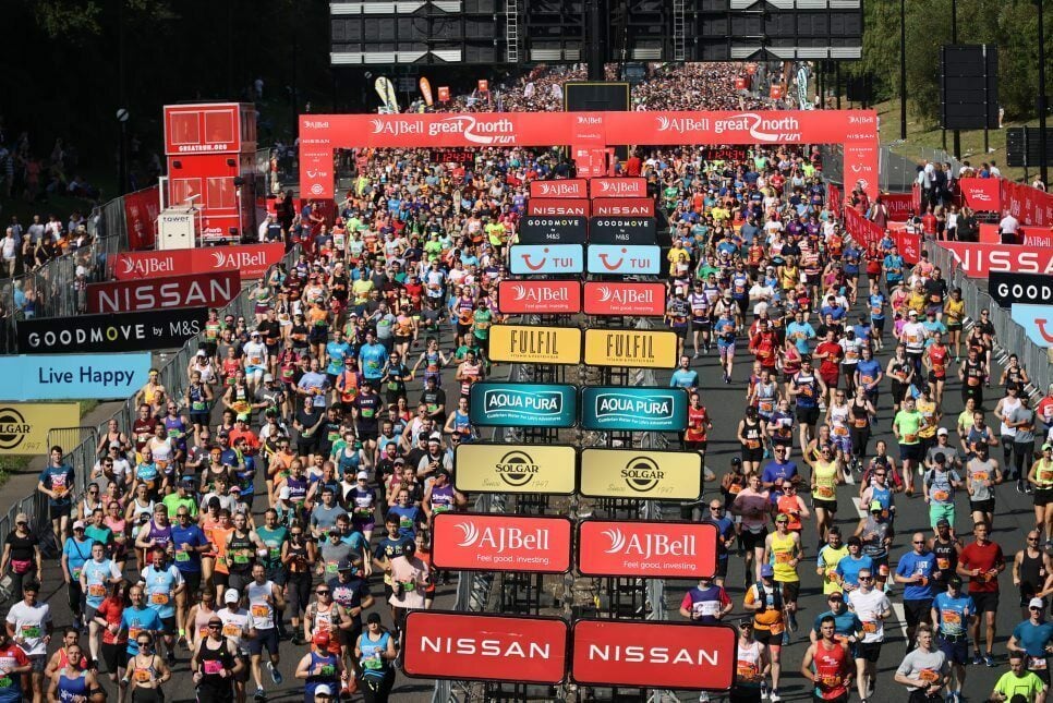 A large crowd of runners participates in the AJ Bell Great North Run, a popular road race. The image shows thousands of runners in colorful athletic gear starting the race under a red starting arch with multiple sponsor logos, including 