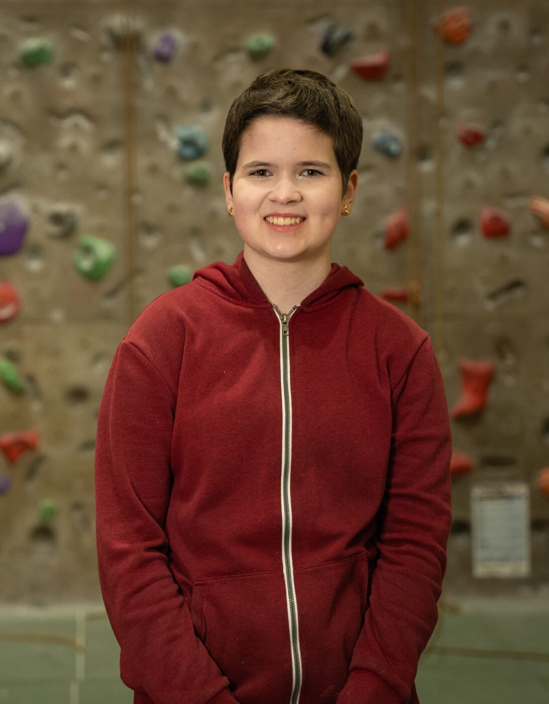 Rosa smiles while standing in front of a climbing wall. They are wearing a red zip-up hoodie and the background features various colorful climbing holds on a textured wall.