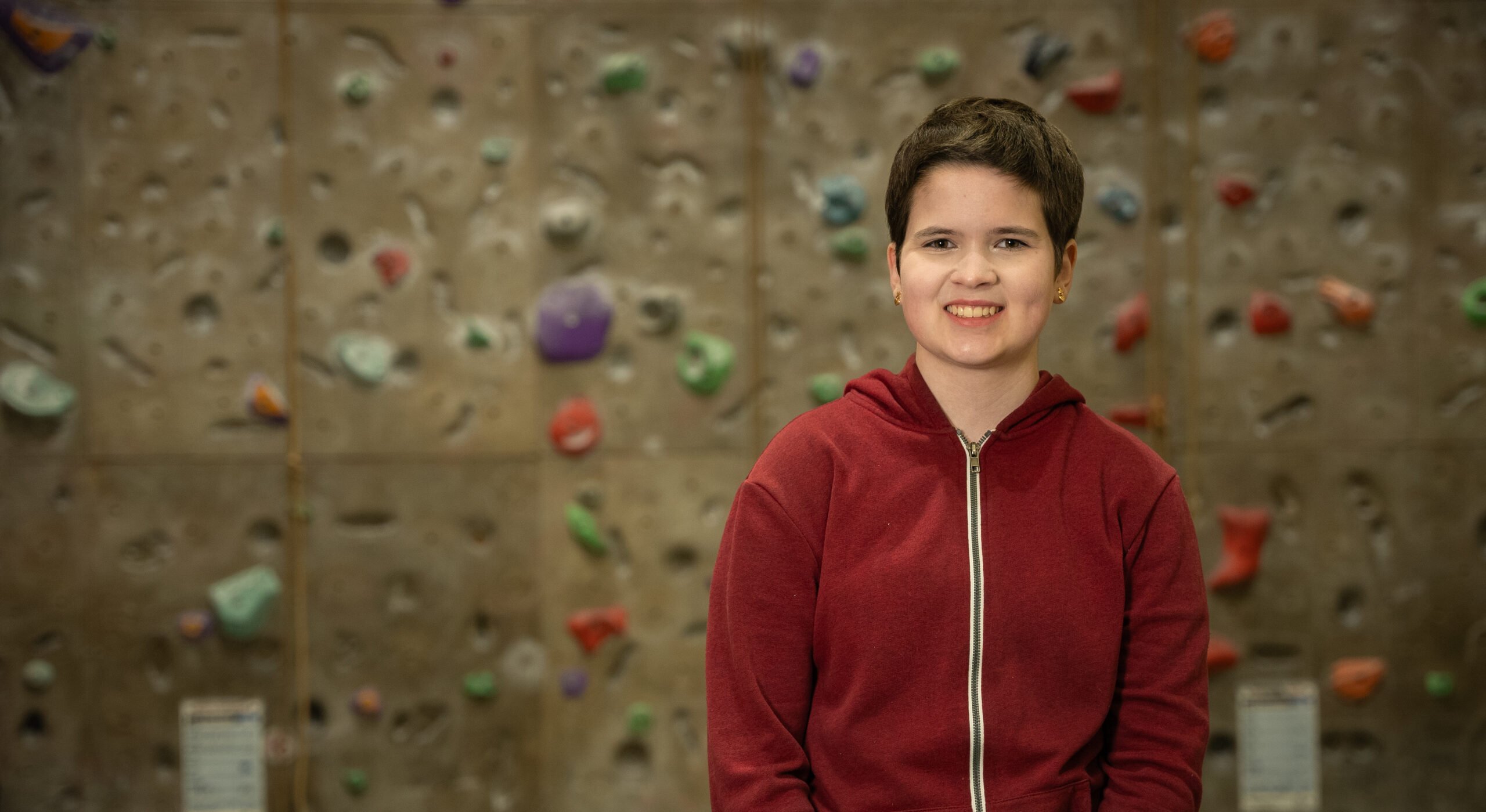 Rosa smiles while standing in front of a climbing wall. They are wearing a red zip-up hoodie and the background features various colorful climbing holds on a textured wall.