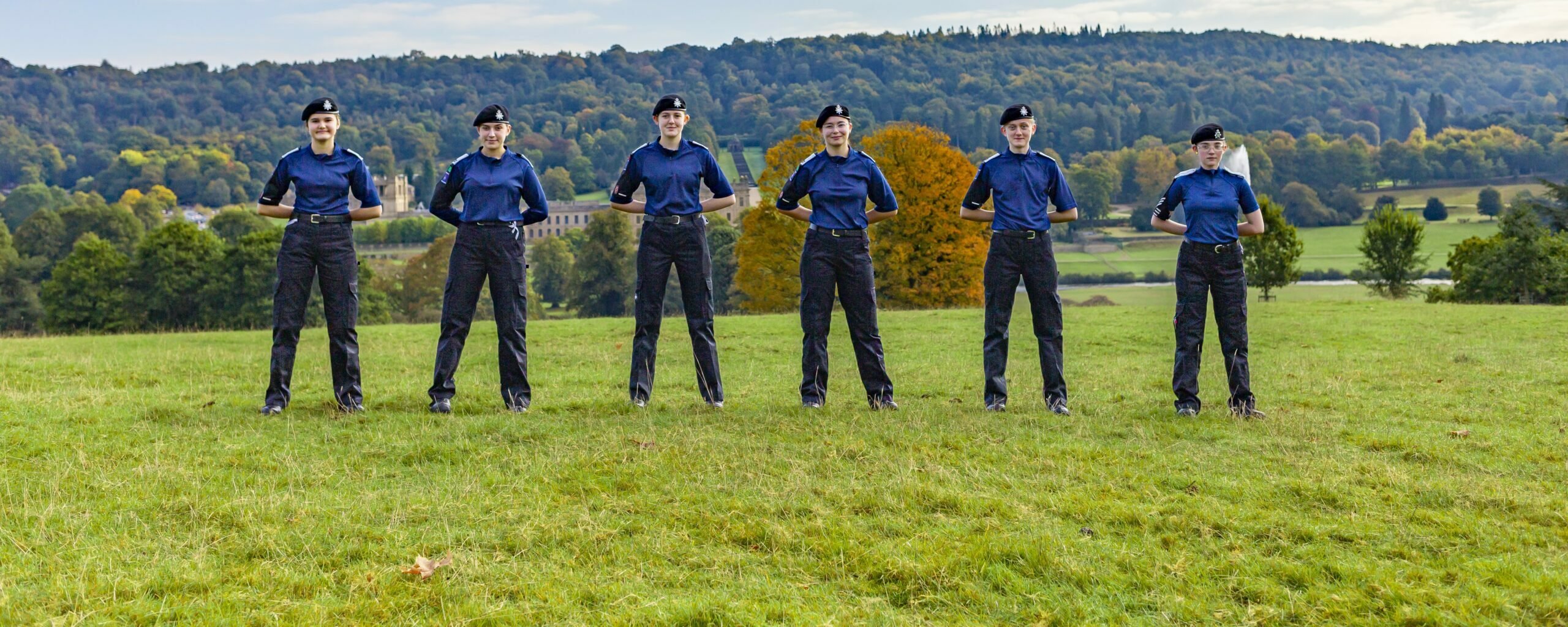 A group of six uniformed individuals stands in a line on a grassy field, facing forward. They are dressed in blue shirts and black pants, with hats on their heads. Behind them, a scenic landscape features rolling hills and trees with autumn foliage under a partly cloudy sky.