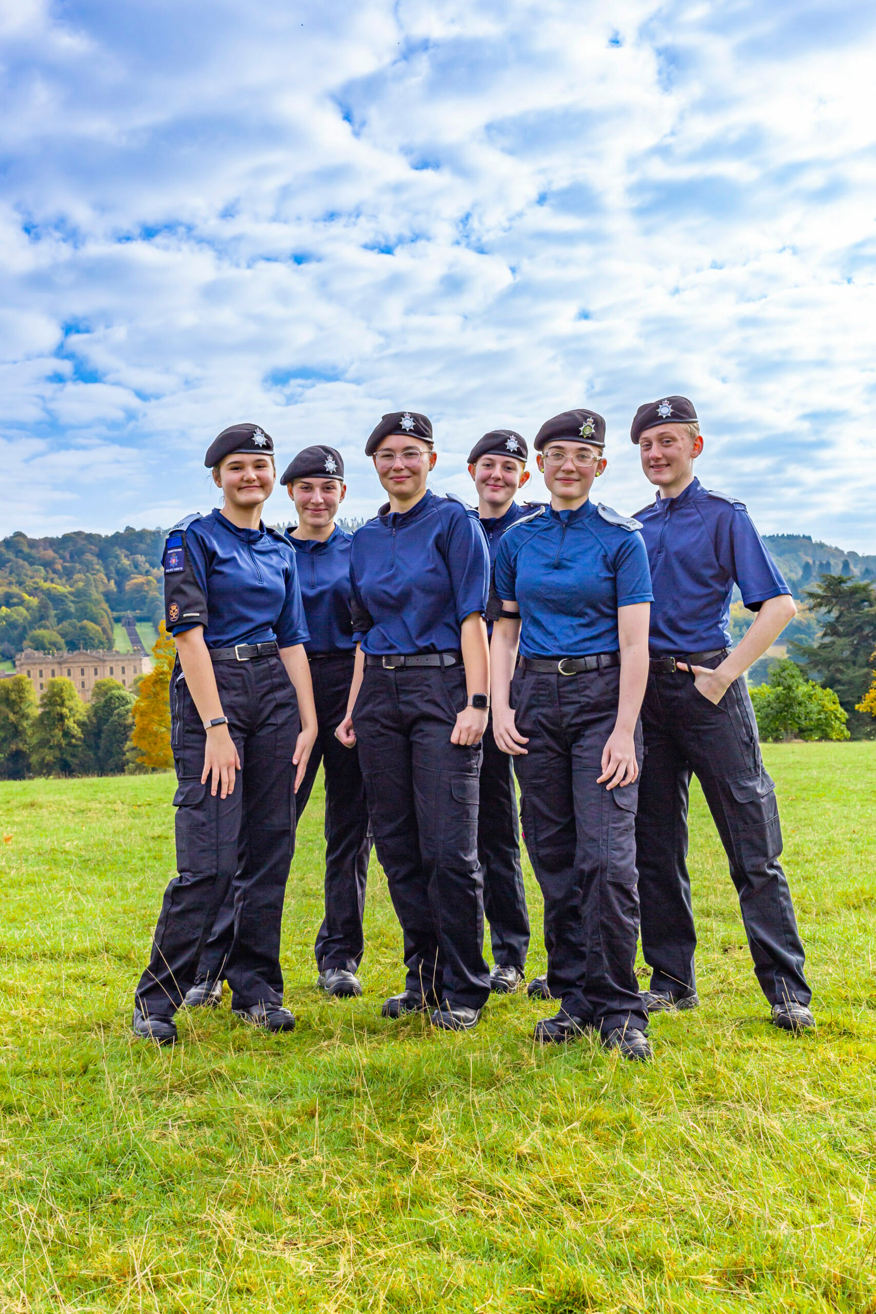 A group of six young individuals dressed in navy blue uniforms and black trousers stands together in a grassy field. They are smiling and posing for the camera, with a scenic landscape of trees and a building in the background under a partly cloudy sky.