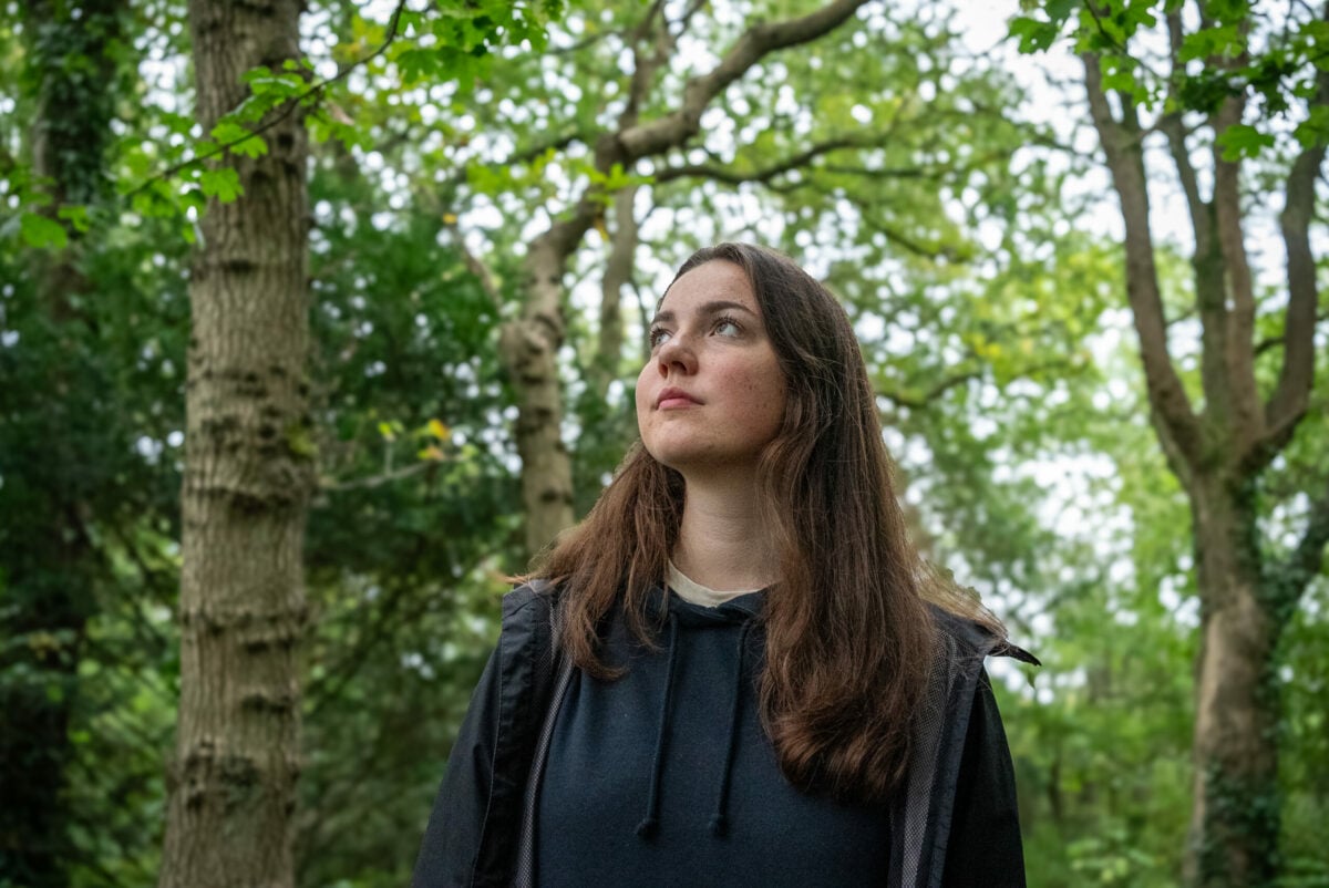 Lila stands in a lush green forest, looking upward with a thoughtful expression. She is wearing a dark hoodie and jacket, surrounded by tall trees with sunlight filtering through the leaves.