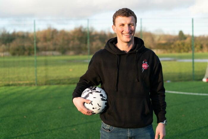 Ryan holding a soccer ball on a green outdoor field with a fence and trees in the background, wearing a black hoodie with a sports-themed logo.