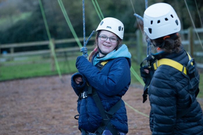 Two young girls are preparing for an outdoor climbing activity. They are wearing helmets and harnesses, with one girl smiling at the camera while the other is looking at her. The background features a grassy area and climbing ropes. The scene is set in a natural environment, suggesting a fun and adventurous atmosphere.