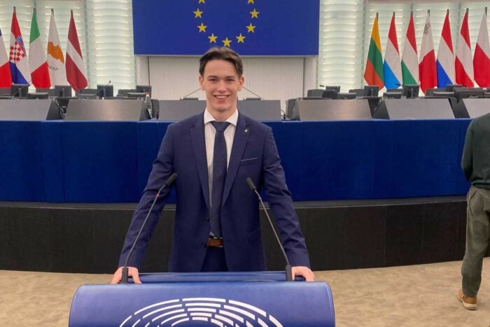 Jake in a dark blue suit, white shirt, and tie stands confidently at a podium in a large conference room. The podium features the emblem of the European Parliament. Behind him are rows of empty desks, microphones, and a blue flag with yellow stars symbolizing the European Union. Several national flags are visible in the background.