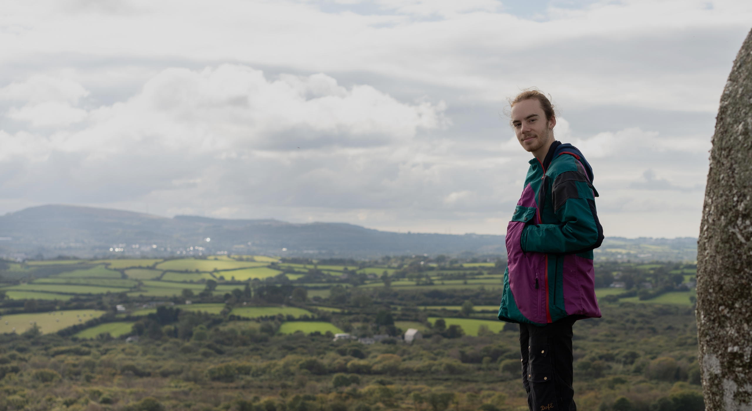 Josh stands on a rocky outcrop overlooking a lush green landscape with rolling hills and fields. They are wearing a colorful jacket and dark pants, with a scenic view of the countryside and cloudy sky in the background.