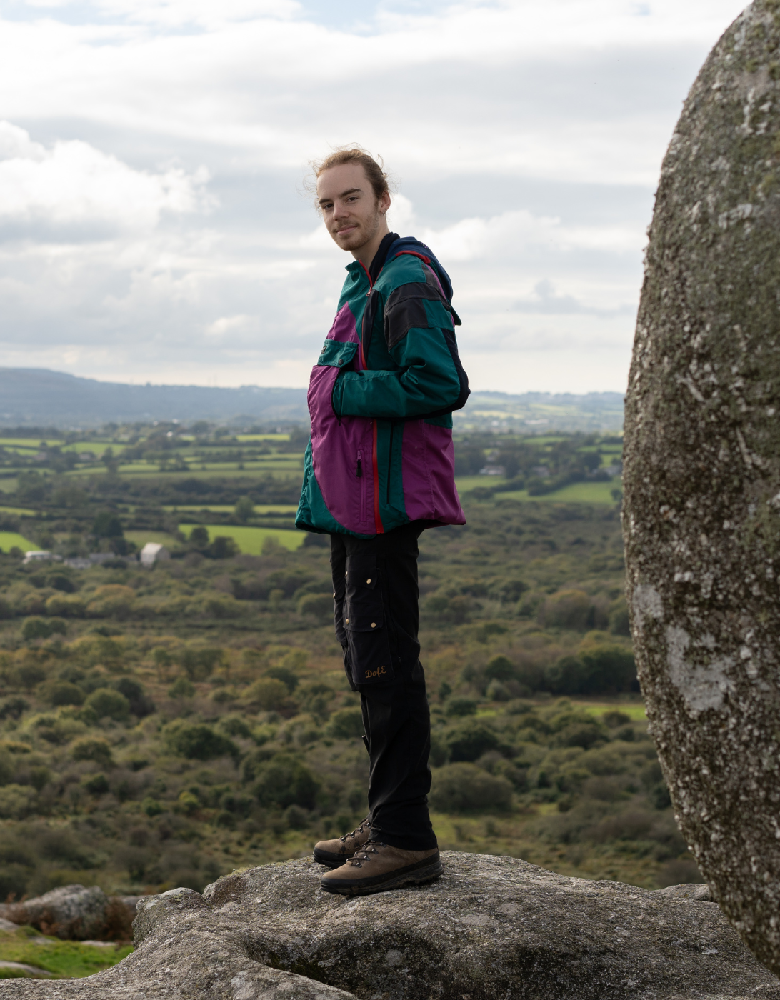 Josh stands on a rocky outcrop overlooking a lush green landscape with rolling hills and fields. They are wearing a colorful jacket and dark pants, with a scenic view of the countryside and cloudy sky in the background.